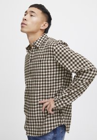 Camisa de patrón de cuadros marrón y blanco con mangas largas, botón en la parte delantera y dos bolsillos en el pecho. Tela suave con un corte casual.