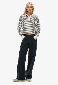 Ung kvinde med langt blond hår har en cropped sort og hvid stribet langærmet polo skjorte, sorte brede bukser og beige sneakers.