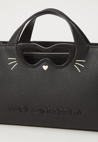 Borsa a tracolla in pelle nera con finitura testurizzata, caratterizzata da dettagli simili a quelli di un gatto e un design unico della maniglia che ricorda gli occhiali da sole. Nome del marchio impresso.