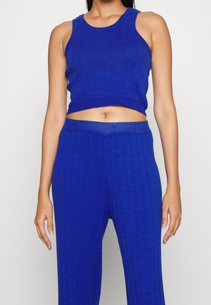 Woman wearing a blue ribbed sleeveless crop top and matching high-waisted pants, standing against a plain light background.