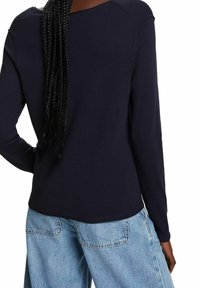 Femme aux longs cheveux tressés portant un haut à manches longues bleu marine et un jean denim bleu clair, vue de dos.
