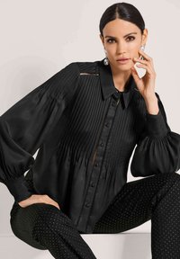 Zwarte geplooide blouse met een kraag, knopen aan de voorkant en volumineuze mouwen. Gecombineerd met zwarte broek met stippenpatroon.