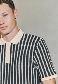 Polo cu mânecă scurtă, prezentând dungi zigzag alternante de culoare bleumarin și crem, cu un guler crem și o închidere cu trei butoane.