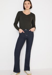 Jeans a zampa di colore blu scuro con cuciture bianche a contrasto, abbinati a una maglia nera aderente a maniche lunghe. La modella è in piedi con le mani nelle tasche.