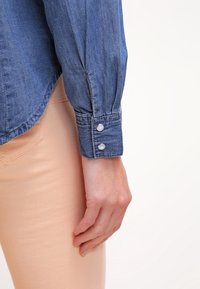 Gros plan sur la main d'une personne reposant le long du corps, portant une chemise en denim bleu avec des poignets à boutons et un pantalon rose clair.