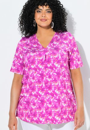 Mujer con cabello negro y rizado, que lleva pendientes de aro con perlas y una blusa de manga corta con estampado floral de color rosa brillante, posando frente a un fondo blanco.
