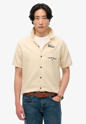 Kortærmet beige skjorte med knaplukning og brystlomme, som har en broderet "West" og et oksehoved-design. Båret med denim jeans og et brunt bælte.