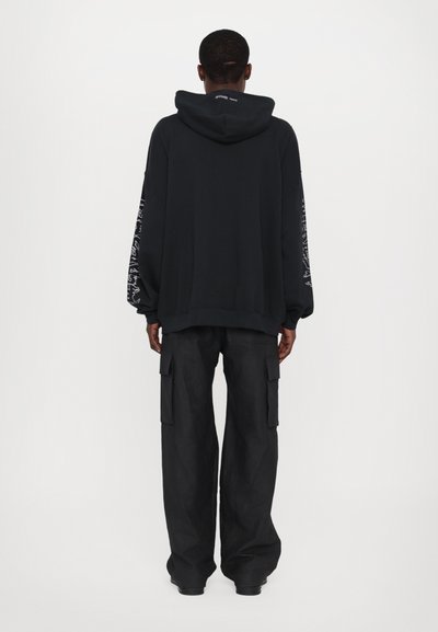 Sudadera negra con capucha, que presenta mangas largas y diseños gráficos blancos en las mangas. Combinada con pantalones cargo negros holgados.