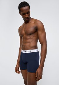 Μυώδης άνδρας που φοράει κοντά εσώρουχα Lacoste σε ναυτικό μπλε με λευκή μέση, στέκεται μπροστά σε ένα απλό ανοιχτό γκρι φόντο.