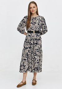 Sukienka midi w kwiatowy wzór w granatowo-kremowych kolorach, z gromadzonym dekoltem, balonowymi rękawami i czarnym paskiem w talii. Brązowe płaskie buty.