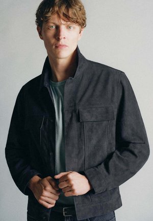 Jeune homme aux cheveux bruns ondulés portant une veste sombre boutonnée sur une chemise verte, regardant droit devant avec une expression neutre.