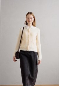 Krämfärgad, texturerad cardigan med krage och knapplås, tillsammans med en lång svart jeanskjol. Mörk handväska över kroppen.