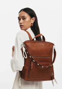 Brauner Leder-Rucksack mit strukturiertem Design, vorderer Reißverschlusstasche, abnehmbarem Tragegurt und geprägt Logo mit metallischen Akzenten.