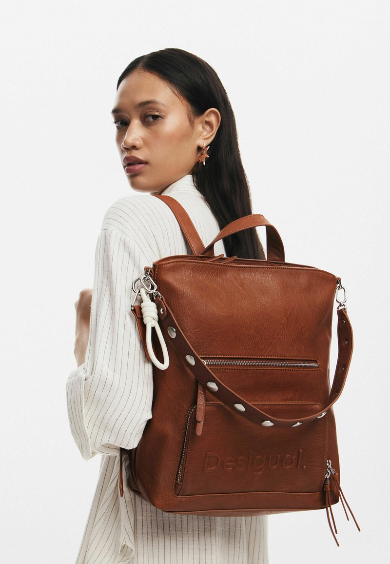 Brauner Leder-Rucksack mit strukturiertem Design, vorderer Reißverschlusstasche, abnehmbarem Tragegurt und geprägt Logo mit metallischen Akzenten.