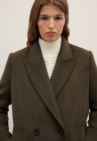 Abrigo cruzado verde oliva con acabado texturizado, solapas con muescas y botones negros, superpuesto sobre un suéter de punto trenzado en color crema.