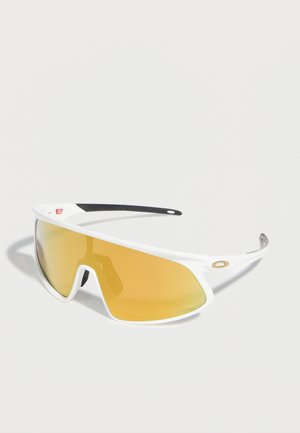 Λευκά αθλητικά γυαλιά ηλίου με περιμετρικούς φακούς με κίτρινη απόχρωση και μαύρες ελαστικές λαβές στους βραχίονες, τοποθετημένα σε ανοιχτή επιφάνεια.