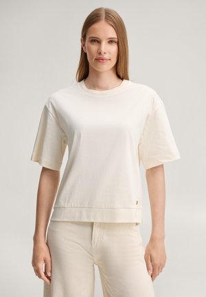 Vrouw met steil lichtbruin haar draagt een offwhite T-shirt met korte mouwen en crèmekleurige broek, staand tegen een effen lichte achtergrond.