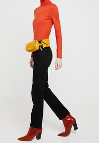 Maglione a collo alto rosso a maniche lunghe, pantaloni aderenti neri, stivali con tacco marroni e una borsa a cintura gialla con un accentuazione con zip.