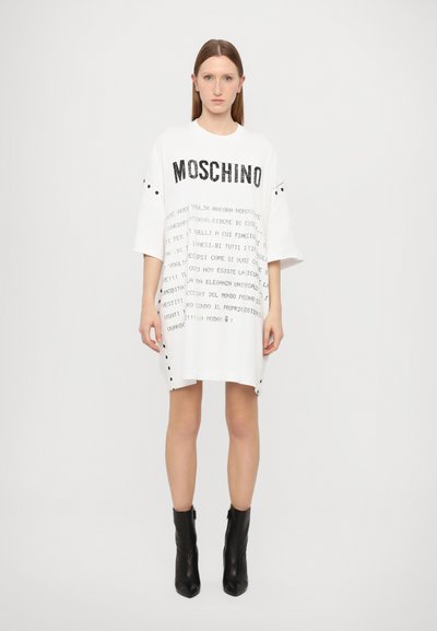 Robe blanche surdimensionnée avec texte imprimé noir, présentant des manches courtes et des détails à pression sur les côtés. Portée avec des bottines noires.