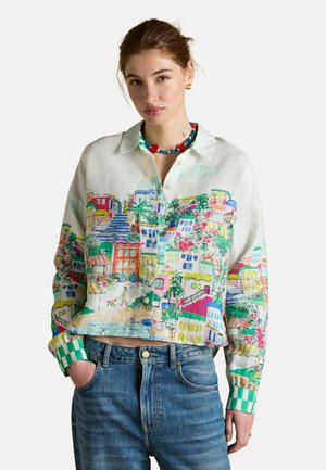 Junge Frau, die ein buntes Hemd mit Druck eines mediterranen Dorfes und blaue Jeans trägt, Hände in den Taschen, neutraler Hintergrund.