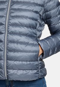 Veste matelassée gonflée, zippée, de couleur bleu clair, avec des coutures horizontales matelassées et des poches latérales. Tissu lisse et brillant avec une texture douce.