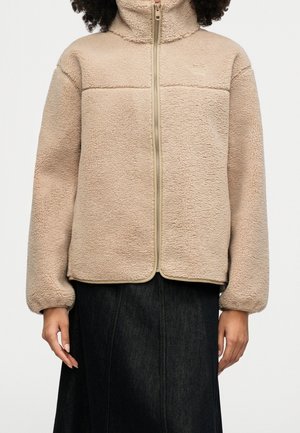 Veste polaire beige zippée avec col haut portée par une personne aux cheveux bouclés et jupe noire, visible du cou aux hanches.