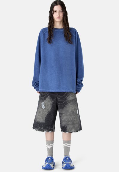 Personne aux longs cheveux foncés portant un sweat-shirt bleu ample, un short noir large usé, des chaussettes grises et des baskets bleues, debout droit.