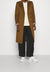 Cappotto doppiopetto in lana marrone, maglione bianco a trecce, pantaloni neri a gamba larga e sneakers bianche chunky. Tessuto liscio e vestibilità su misura.