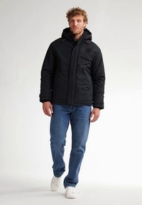 Jachetă izolată neagră cu glugă, închidere cu fermoar și detalii cu buzunare, combinată cu blugi albaștri și adidași albi. Îmbrăcăminte casual pentru exterior.