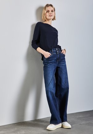 Dunkelblaue weite Jeans mit hoher Taille und frontalen Taschen, kombiniert mit einem taillierten marineblauen Oberteil und weißen Sneakers auf einem Betonboden.