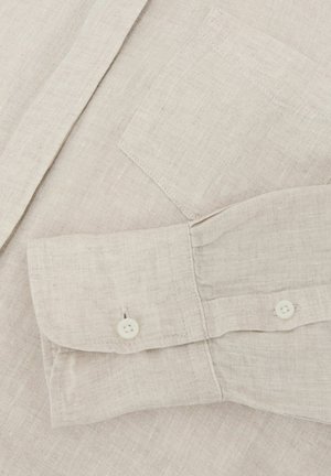 Manica di camicia in lino beige chiaro con polsino abbottonato e taschino sul petto, che mostra la trama del tessuto e i dettagli delle cuciture.