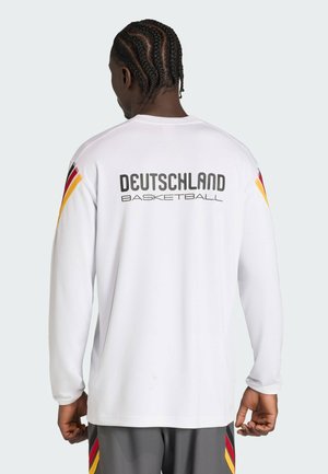 Personne aux cheveux tressés portant une chemise blanche à manches longues avec le texte "Deutschland Basketball" et les couleurs du drapeau allemand sur les manches et le short.