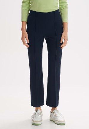 Marineblaue maßgeschneiderte Hose mit geradem Bein, strukturiertem Stoff und sichtbaren Faltendetails. Kombiniert mit weißen Sneakers mit grünen Akzenten.