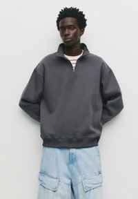 Grauer Zip-Hoodie mit hohem Kragen, lässiger Passform und geripptem Saum. Kombiniert mit hellblauen Cargo-Hosen mit großen Taschen.