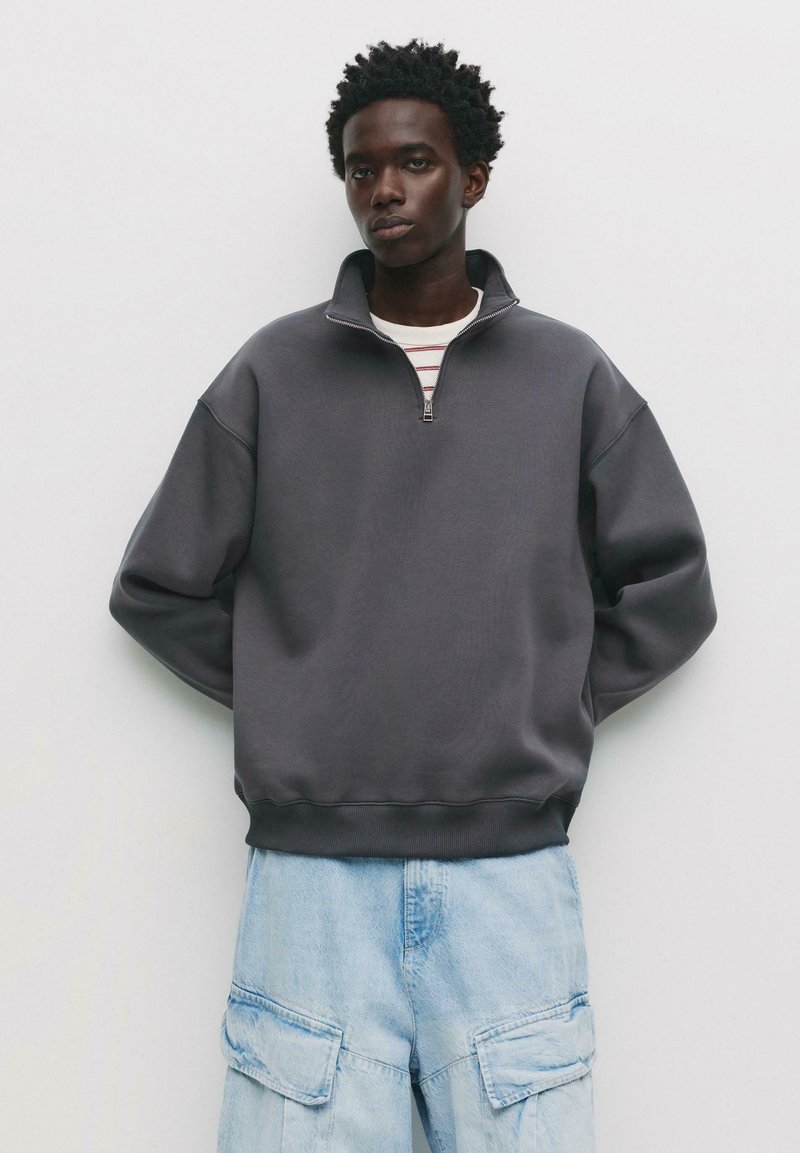 Grauer Zip-Hoodie mit hohem Kragen, lässiger Passform und geripptem Saum. Kombiniert mit hellblauen Cargo-Hosen mit großen Taschen.