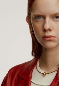 Chaqueta de cuero roja, blusa blanca, collar de oro con un colgante redondo texturizado. Presenta un primer plano de un rostro con ojos azules y cabello rojo claro.