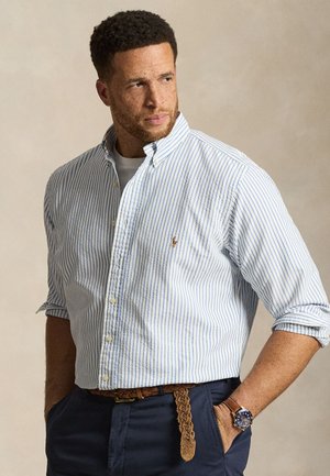 Homme aux cheveux courts et bouclés portant une chemise blanche à rayures bleues boutonnée, un pantalon bleu marine, une ceinture marron tressée et une montre-bracelet, regardant vers la gauche.