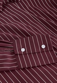 Bordó csíkos ing vékony fehér csíkokkal, két fehér gombbal az ujjatlan mandzsettán, és sima szövetes, normál gallérral.