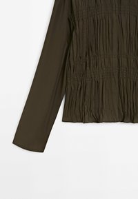 Blouse vert olive foncé à manches longues avec des détails en tissu froncé et texturé sur le devant et des manches lisses.