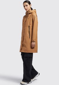 Beige wasserfester langer Mantel mit Kapuze, Frontreißverschluss und seitlichen Taschen. Mit schwarzen Akzenten und einer lockeren Passform kombiniert mit schwarzen Cargo-Hosen.