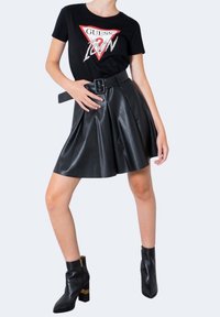 T-shirt noire avec logo "GUESS", détail graphique et manches courtes, associée à une jupe plissée en similicuir noir et des bottines à la cheville.