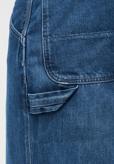 Κοντινό πλάνο μπλε τζιν παντελονιού που δείχνει την πίσω τσέπη και τη θηλιά εργαλείων με ορατή ραφή και υφή υφάσματος.