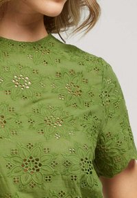 Haut brodé vert présentant des motifs floraux et des découpes. Texture lisse, manches courtes et encolure ronde.