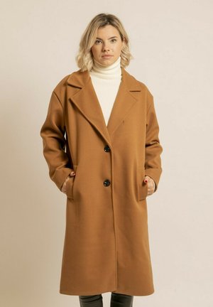 Femme debout, les mains dans les poches, portant un long manteau camel sur un col roulé blanc, sur un fond uni.