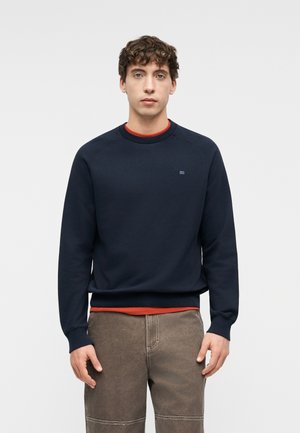 Fiatal férfi, aki előre néz, sötétkék pulóvert visel narancssárga ing felett, barna nadrágban, egyszínű világos háttér előtt.