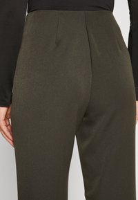 Höga olivgröna byxor tillverkade av ett slätt, strukturerat tyg med veck på baksidan och en åtsittande midja.