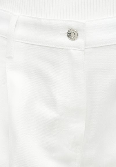 Κοντινό πλάνο στη μέση λευκών τζιν denim με ασημί μεταλλικό κουμπί και θηλιά για ζώνη, φορεμένα πάνω από λευκό ριμπ ύφασμα τοπ.