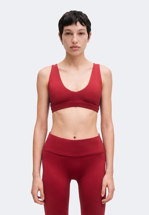 Kvinde med kort mørkt hår står fremadvendt iført matchende rød sports-bh og højtaljede leggings mod en lys ensfarvet baggrund.