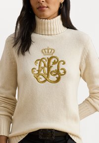 Femeie îmbrăcată într-un pulover crem cu guler înalt, având monogramă brodată cu fir aurit și detalii cu coroană, combinat cu brâu și pantaloni negri.