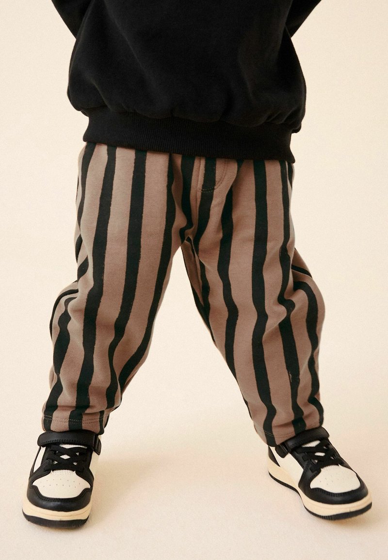 Enfant portant un pantalon marron avec des rayures verticales noires et des baskets noires et blanches, debout jambes écartées sur un fond clair.